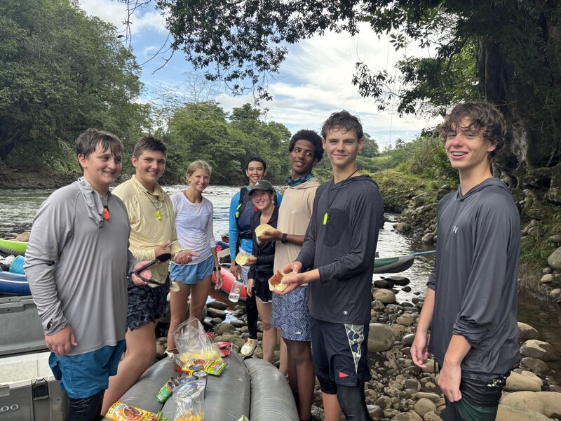 A group of young people are standing near a river, possibly after a rafting or kayaking trip. They appear to be taking a break, with some holding snacks. The background features lush greenery and trees, suggesting a natural, outdoor setting. The atmosphere seems relaxed and friendly, as they are all smiling and looking at the camera.